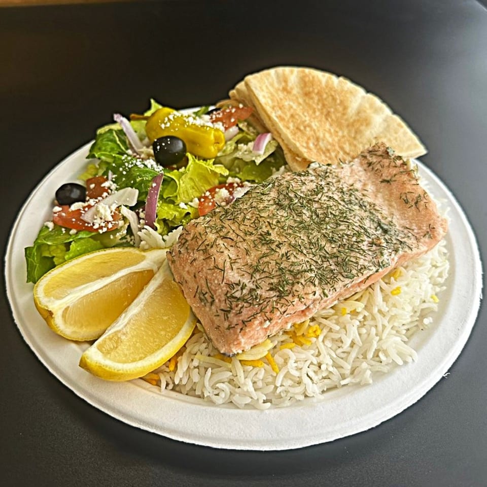 Mediterranean salmon with dill, rice, lemon wedges, Greek salad, and pita bread on a white plate.