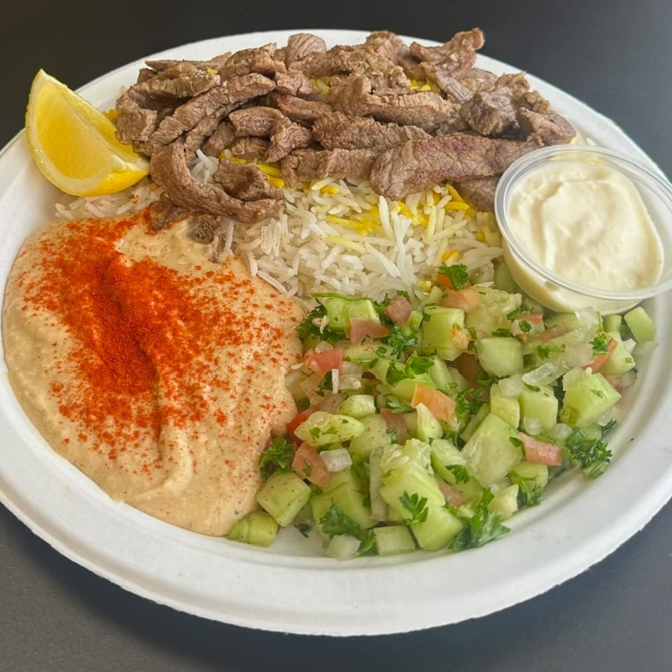 Mediterranean platter with sliced beef, rice, hummus, cucumber salad, and a lemon wedge.